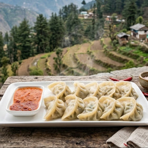 A white rectangular plate of steamed dumplings with a side of red dipping sauce, set on a rustic wooden table with a mountain village and terraced hills in the background.