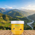 Yellow jar of Mamaearth cream on a wooden surface with a scenic mountain landscape in the background
