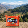 Samyang Ramen package on a wooden surface with a scenic mountain landscape in the background