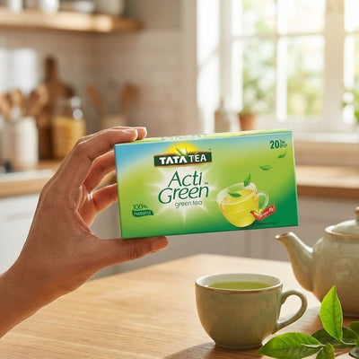 Hand holding a box of Tata Tea Acti Green tea with a cup and teapot on a table.